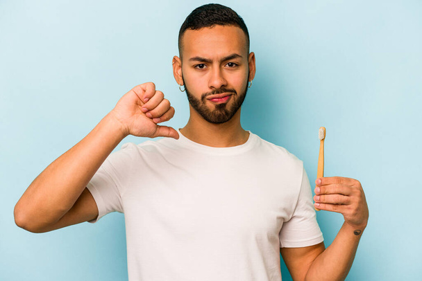 Young hispanic man brushing teeth isolated on blue background showing a dislike gesture, thumbs down. Disagreement concept. - Foto, afbeelding