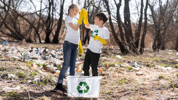 Αγόρι και κορίτσι με ένα κοντέινερ σε μια συλλογή πλαστικών σκουπιδιών σε ένα μολυσμένο ξέφωτο, με πινακίδες ανακύκλωσης στα μπλουζάκια - Φωτογραφία, εικόνα