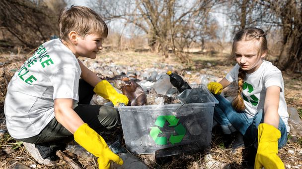 Αγόρι και κορίτσι μαζεύουν πλαστικά σκουπίδια σε ένα κοντέινερ σε ένα μολυσμένο ξέφωτο, ανακυκλώνοντας πινακίδες στα μπλουζάκια. - Φωτογραφία, εικόνα