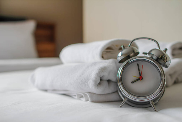 目覚まし時計はお客様がリゾートに滞在し、タオルとしてマットレスの上に配置するために折り畳まれている白いスパタオルの隣に座っている彼らは休息する前にシャワーを取る必要があるとき. - 写真・画像