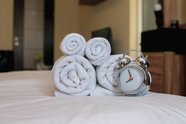Wecker sitzt neben sauberen weißen Wellness-Handtüchern, die für Gäste des Resorts zusammengefaltet und auf ihre Matratzen gelegt werden, da Handtücher ein Muss sind, wenn sie vor der Ruhe duschen müssen.. - Foto, Bild
