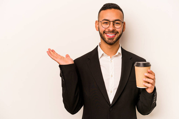 Junger hispanischer Geschäftsmann mit Kaffee zum Mitnehmen auf weißem Hintergrund, der einen Kopierraum auf einer Handfläche zeigt und eine andere Hand auf der Taille hält. - Foto, Bild