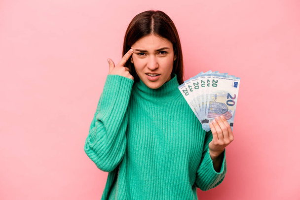 Young caucasian woman holding banknotes isolated on pink background showing a disappointment gesture with forefinger. - Foto, Imagen