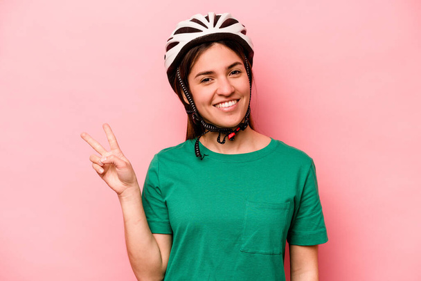 ピンクの背景に隔離されたヘルメットを身に着けている若い白人女性喜びと屈託のない指で平和のシンボルを示す. - 写真・画像