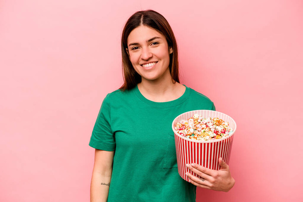 jonge Kaukasische vrouw houden popcorn geïsoleerd op roze achtergrond gelukkig, glimlachend en vrolijk. - Foto, afbeelding