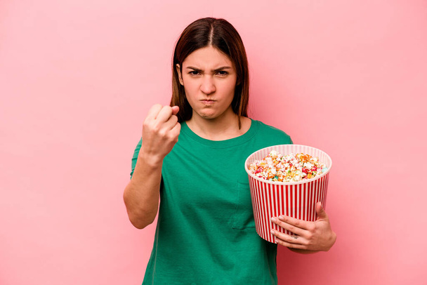 Young caucasian woman holding popcorn isolated on pink background showing fist to camera, aggressive facial expression. - Photo, image