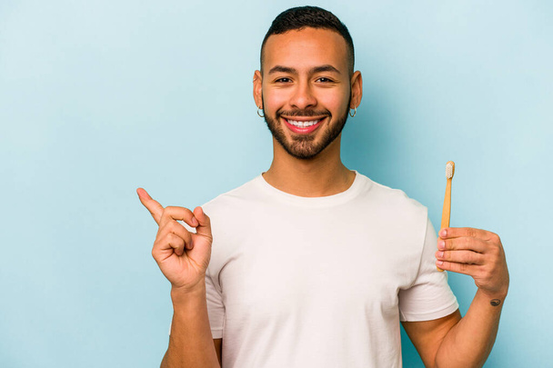 Young hispanic man brushing teeth isolated on blue background smiling and pointing aside, showing something at blank space. - Foto, Imagen