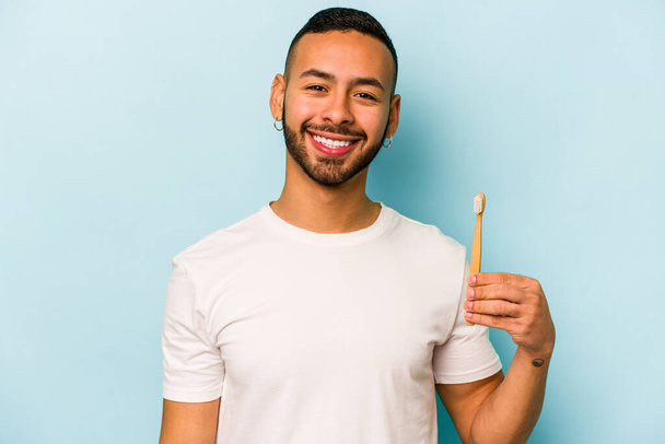 青の背景に孤立した若いヒスパニック系の男の歯をブラッシング幸せな笑顔と陽気. - 写真・画像