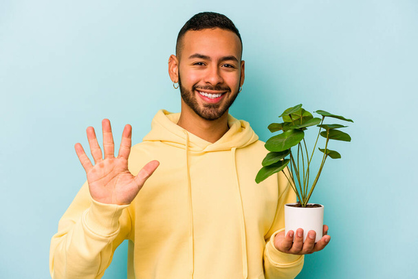Joven hispano sosteniendo una planta aislada sobre fondo azul sonriente alegre mostrando el número cinco con los dedos. - Foto, Imagen