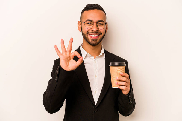 Joven empresario hispano sosteniendo café para llevar aislado sobre fondo blanco alegre y confiado mostrando gesto ok. - Foto, Imagen