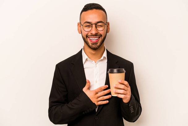 Junger hispanischer Geschäftsmann mit Imbisskaffee auf weißem Hintergrund lacht laut aus und hält die Hand auf der Brust. - Foto, Bild