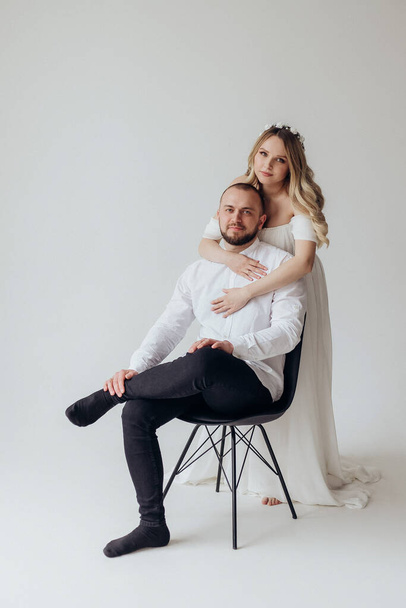 Husband and pregnant blonde wife in a white dress in the studio on a white background with a wreath on their heads hold their hands on their stomachs - Foto, Bild