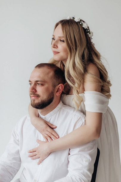 Husband and pregnant blonde wife in a white dress in the studio on a white background with a wreath on their heads hold their hands on their stomachs - Foto, Imagem