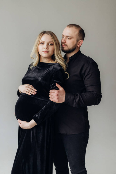 Husband and pregnant blonde wife in a black dress in the studio on a black background stand and hug - Zdjęcie, obraz