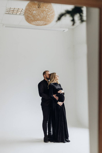 Husband and pregnant blonde wife in a black dress in the studio on a black background stand and hug - Foto, Imagem