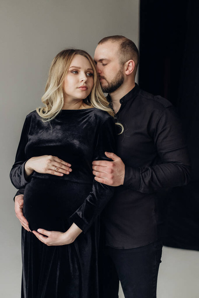 Férj és terhes szőke feleség fekete ruhában a stúdióban egy fekete háttér stand és ölelés - Fotó, kép