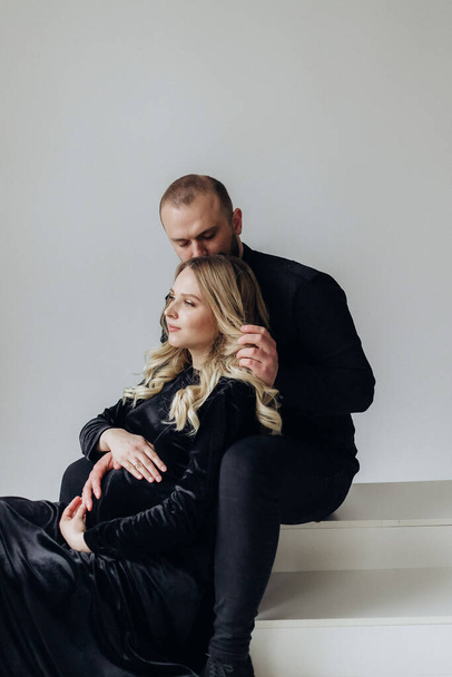 Husband and pregnant blonde wife in a black dress in the studio on a black background sitting and hug - Foto, Imagem