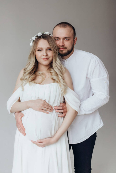 Manžel a těhotná blondýna manželka v bílých šatech v ateliéru na bílém pozadí s věncem na hlavě držet ruce na břiše - Fotografie, Obrázek
