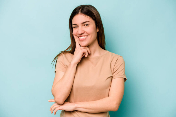 Young caucasian woman isolated on blue background smiling happy and confident, touching chin with hand. - Photo, Image
