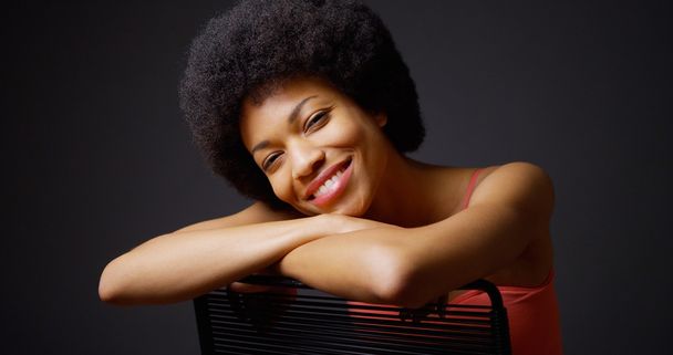 Χαμογελώντας μαύρη γυναίκα που κάθεται με τα χέρια πάνω από πίσω από καρέκλα - Φωτογραφία, εικόνα