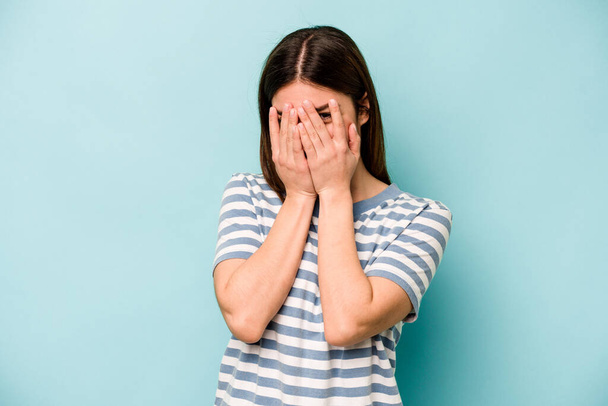 Young caucasian woman isolated on blue background blink at the camera through fingers, embarrassed covering face. - Foto, Imagen