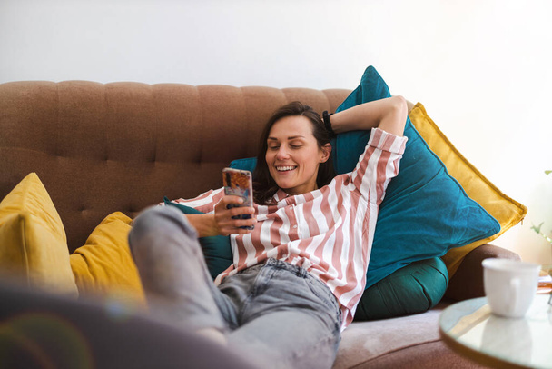 Szczęśliwa młoda kobieta za pomocą telefonu komórkowego na kanapie - Zdjęcie, obraz