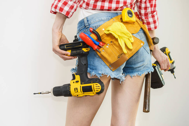 Woman wearing protective workwear toolbelt using drill. Girl working at flat remodeling. Building, repair and renovation. - Фото, изображение