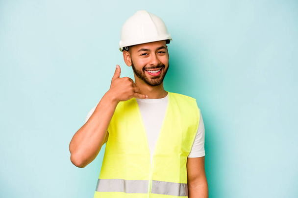 Mladý dělník hispánec izolovaný na modrém pozadí ukazující gesto mobilního telefonu s prsty. - Fotografie, Obrázek