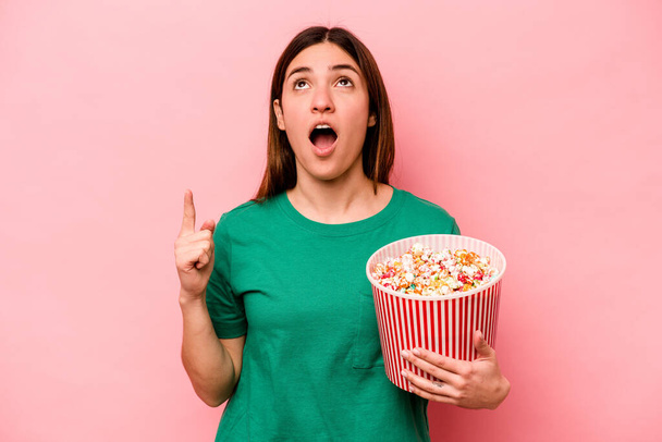 Young caucasian woman holding popcorn isolated on pink background pointing upside with opened mouth. - Foto, Bild