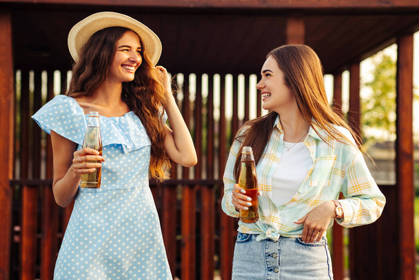 Two young beautiful smiling women in summer clothes and hats. Carefree women have a picnic on the street. Positive girls talking drinking drinks, outdoors in the park in nature - Fotó, kép