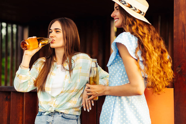 Two young beautiful smiling women in summer clothes and hats. Carefree women have a picnic on the street. Positive girls talking drinking drinks, outdoors in the park in nature - Photo, image