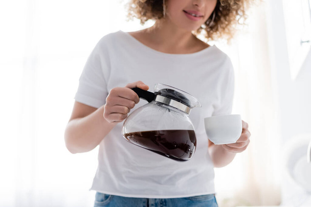 bijgesneden weergave van krullende vrouw in wit t-shirt gieten ochtend koffie - Foto, afbeelding