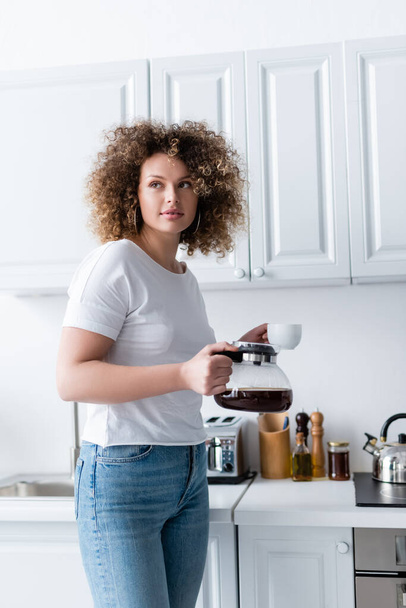 curly woman in white t-shirt standing with cup and coffee pot in kitchen - Photo, image