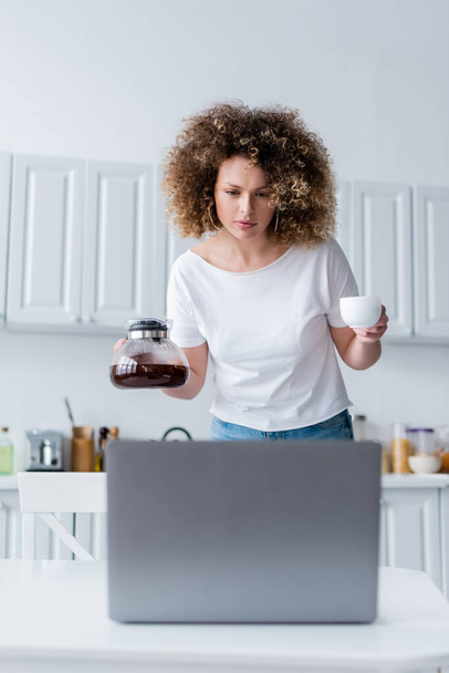 young woman holding cup and coffee pot while looking at laptop in kitchen - Photo, Image