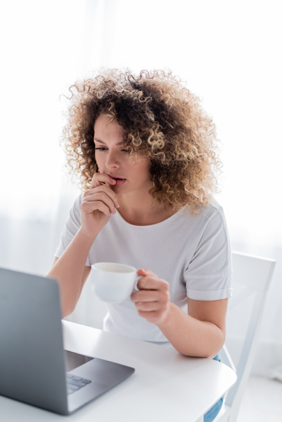 gondolkodó nő csésze kávé gondolkodás közelében laptop a konyhában - Fotó, kép