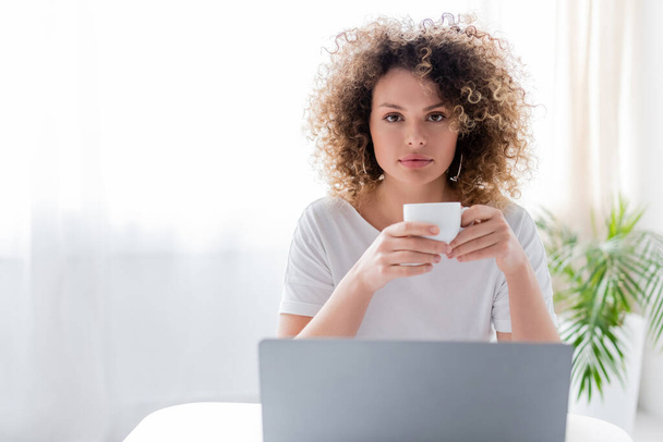 mujer bonita y rizada sosteniendo la taza de café y mirando a la cámara cerca de la computadora portátil - Foto, Imagen
