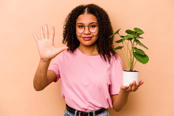 Jovem afro-americana segurando uma planta isolada no fundo bege sorrindo alegre mostrando número cinco com os dedos. - Foto, Imagem
