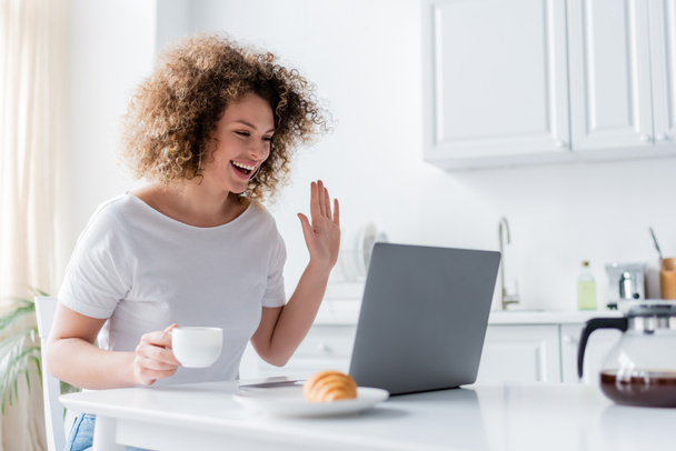 happy woman with coffee cup waving hand near laptop and blurred croissant - Photo, Image