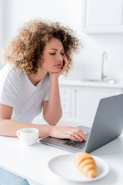 donna pensosa con mano vicino al viso utilizzando il computer portatile vicino a tazza di caffè - Foto, immagini