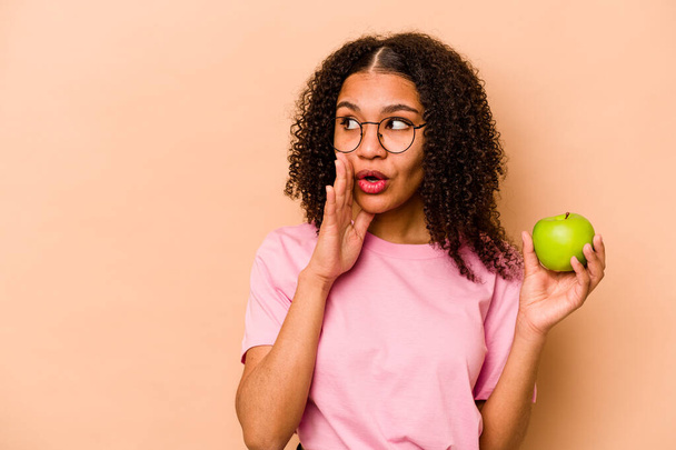 Young African American woman holding an apple isolated on beige background is saying a secret hot braking news and looking aside - Zdjęcie, obraz