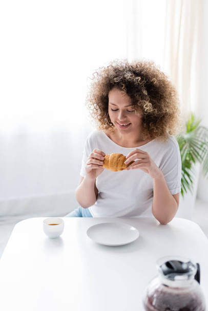 smiling woman with delicious croissant near cup and burred coffee pot - Foto, Imagen