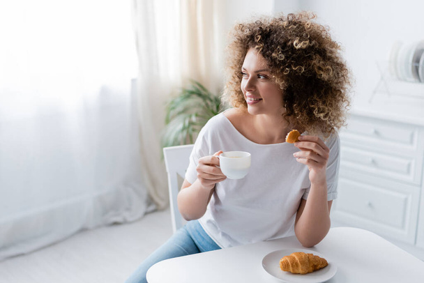 happy woman with croissant and cup of coffee looking away in kitchen - Foto, afbeelding