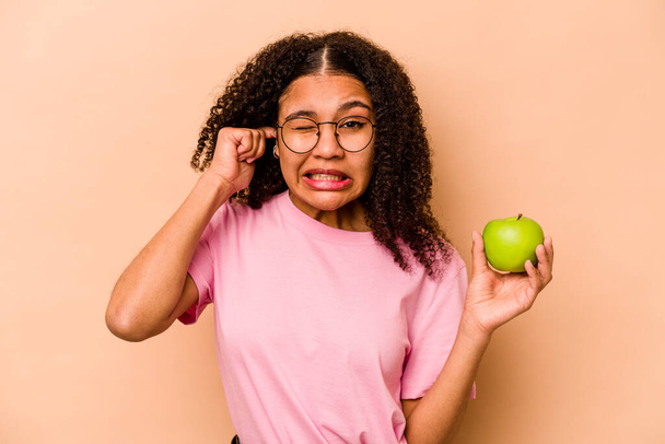 Young African American woman holding an apple isolated on beige background covering ears with hands. - Photo, image
