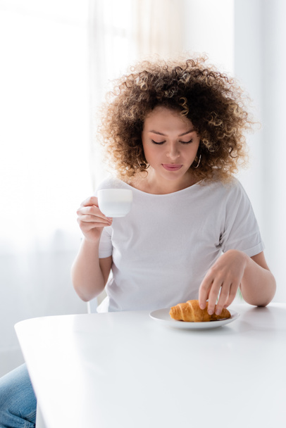 curly woman in white t-shirt holding cup of coffee near tasty croissant - Fotoğraf, Görsel