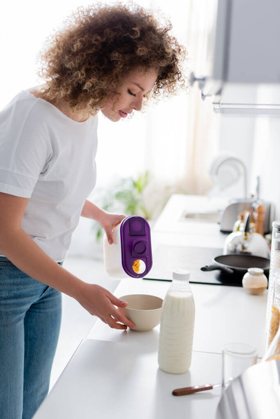 curly woman holding container with corn flakes near bowl and bottle of milk - Фото, зображення