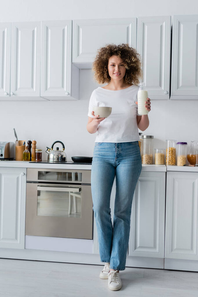 полная длина кудрявая женщина в джинсах держа свежее молоко и миску на кухне - Фото, изображение
