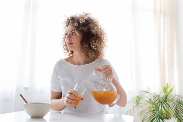 mulher com cabelo ondulado derramando suco de laranja perto da tigela na mesa - Foto, Imagem