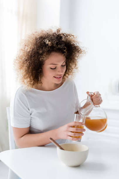 young and positive woman with wavy hair pouring orange juice from jug into glass - Zdjęcie, obraz