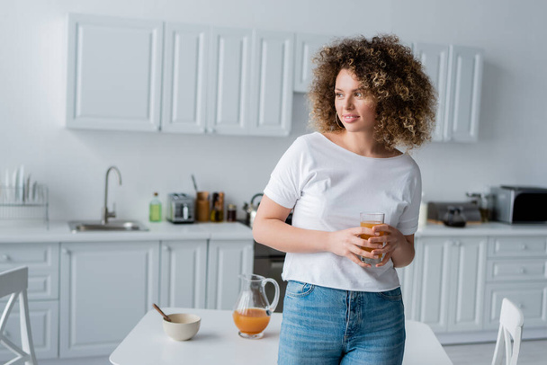 curly woman with glass of fresh orange juice smiling in blurred kitchen - Zdjęcie, obraz