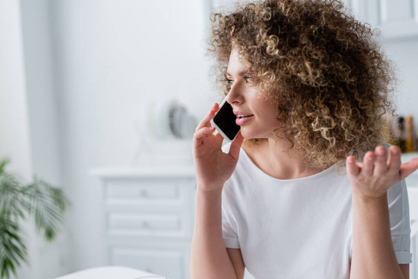 curly woman in white t-shirt gesturing while talking on smartphone at home - Foto, immagini